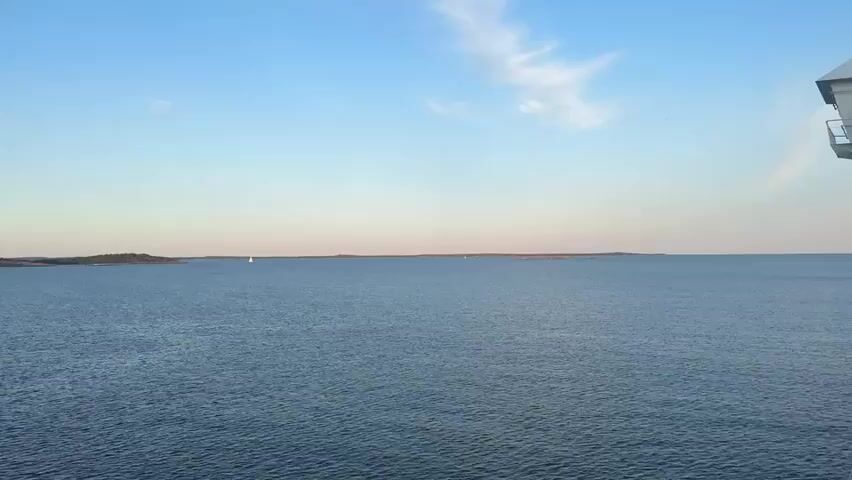 A wide shot shows a vast expanse of calm, blue water under a clear, pale blue sky with a few wispy white clouds. In the distance, on the left side of the frame, a small white sailboat is visible. Further to the right, there are several islands covered in dark green trees and rocky terrain. The sun appears to be low in the sky, casting a warm glow, possibly during sunrise or sunset. The right edge of the frame shows a small portion of a white structure with a railing, suggesting the viewpoint is from a boat or balcony. The water has gentle ripples, indicating minimal wind. The overall scene is serene and natural.