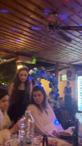 The video shows a gathering at an outdoor venue with a wooden ceiling. Colorful lights are projected onto the ceiling, creating a festive atmosphere. In the foreground, several young women are seated at a table. One woman on the left wears a white fur coat and appears to be looking down at something. Another woman in the center wears a black dress and a tiara, and she is waving. A third woman to the right wears a white top and looks towards the camera. Behind them, a balloon arch in blue and white is visible, along with a woman in the background standing. The scene suggests a celebration, possibly a birthday or a formal event.