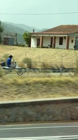 A person wearing a blue shirt and a helmet rides a white scooter on a dirt road. Behind the scooter, a small house with a red roof is visible. In the background, a mountain range can be seen. The scene appears to be in a rural area with dry vegetation and a few trees. The scooter is moving from left to right across the frame. The footage is shaky, suggesting it was filmed from a moving vehicle.