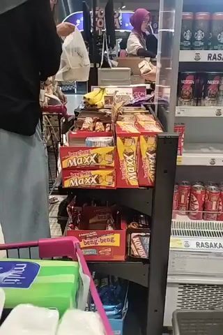 The video shows a person in a black shirt and light blue pants standing in front of a shopping cart, holding a white plastic bag and reaching for items on a display shelf. Behind them, another person wearing a pink hijab is at a checkout counter. The shelves are filled with various snack items, including boxes of "Maxx" and "beng-beng" chocolate bars, as well as cans of drinks like Nescafe and Kit Kat in a refrigerated display. The environment appears to be a supermarket aisle.