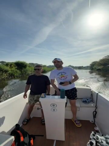 Two men are on a boat, cruising on a narrow river or canal under a sunny sky.  The man on the right is at the helm, holding his phone and dancing while the boat moves forward. He wears a white Converse T-shirt, black Nike shorts, a white baseball cap, and yellow flip-flops. The man on the left wears a black T-shirt, camouflage shorts, sunglasses, and dark flip-flops, and is pointing to the side. A blue and green portable speaker and a beer can are placed on the helm. A third person, a woman, sits on the back seat of the boat wearing sunglasses and giving a peace sign, with a can of soda in her hand. Her backpack is on the seat next to her.  The surroundings include lush green vegetation along the riverbanks. The boat is white with a wooden floor. The camera angle is low, looking up.