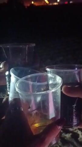 The video begins with a dark, blurry foreground of what appears to be cups on a surface. The background is very dark, with streaks of light suggesting movement or distant lights. As the video progresses, the background becomes clearer, revealing a dark sky with bursts of fireworks. The fireworks are colorful, with shades of pink, white, and yellow visible. There are also faint lights along a distant horizon, possibly indicating a town or cityscape. The foreground remains largely out of focus and dark throughout the clip. There is no clear interaction or identifiable people visible. The overall setting appears to be outdoors at night, likely a beach or open area, with a fireworks display in progress.