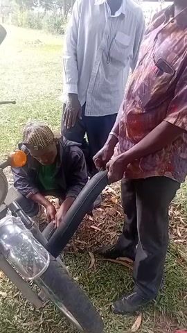 Three people stand outdoors in a grassy area beside a parked motorcycle. One person is crouched down working on something near the front tire of the motorcycle, while two other people stand behind observing. The person crouching is wearing a dark jacket and head covering. One standing person wears a white shirt and dark pants. The other standing person wears a patterned button-down shirt and dark pants. The motorcycle is silver with an orange turn signal. The person crouching appears to be handling a long black object. All three appear to be conversing, though audio is not available.