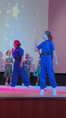 A group of young girls is performing a dance routine on a stage. The girls are dressed in matching blue jumpsuits with red accents, and some are wearing red bows in their hair. The stage backdrop is a large screen displaying a pattern of stars. The lighting is colorful, with hues of pink, purple, and yellow. The girl in the foreground on the right is wearing a blue jumpsuit and white sneakers, and she is dancing with her arms extended. Another girl in a similar blue jumpsuit is on the left, also dancing. Behind them, three girls in floral print dresses with white sashes are standing and singing. The girls appear to be enjoying themselves. The overall scene suggests a children's performance or recital.