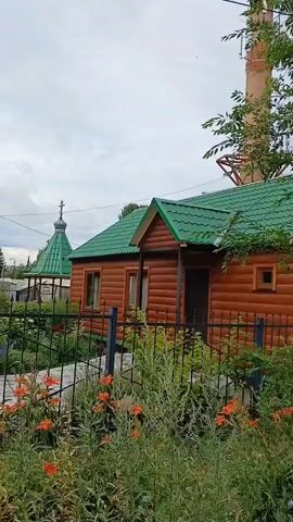 This media file captures an outdoor view of a small, wooden church complex in Volsk, Russia. The primary subjects are the wooden buildings, which appear to be religious structures, possibly a main church and smaller chapels or ancillary buildings. These structures are characterized by their warm, brown wooden exteriors and vibrant green roofs. One building features a distinct golden dome topped with a cross, a common architectural element in Orthodox churches.

The scene is set within a well-maintained courtyard, enclosed by a black metal fence. Beyond the fence, lush greenery and flowering plants, including bright orange lilies, add color and life to the foreground. A paved walkway or courtyard area is visible within the fenced enclosure. Tall, mature trees with dense green foliage flank the buildings, suggesting a natural or park-like setting. In the background, glimpses of other buildings and utility poles indicate a semi-urban or rural residential area.

The weather appears to be overcast, with a cloudy sky dominating the upper portion of the frame. The lighting suggests it is daytime, likely late morning or afternoon, given the brightness despite the clouds. No people are visible in the media file, and the primary activity appears to be the static depiction of the church complex and its surroundings. The overall impression is one of tranquility and perhaps a sense of community or spiritual importance associated with the location. The wooden construction and the presence of a church with a golden dome are strong indicators of Russian Orthodox architecture and religious practice.