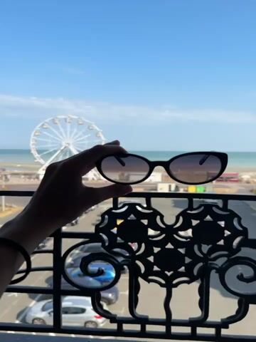 The video opens with a close-up shot of a hand holding up a pair of black sunglasses against a bright blue sky and the ocean. The sunglasses have a dark frame and tinted lenses. In the background, a large white Ferris wheel is visible, along with a beach and some buildings. The camera then pans down slightly, revealing a black metal railing with an ornate design in the foreground. Below the railing, a parking lot filled with cars can be seen. The scene appears to be a sunny day at a seaside resort. The video is a short loop, with the slight movement of the sunglasses and a subtle shift in lighting being the only apparent changes.