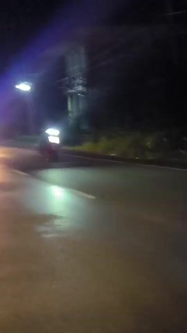 The video shows a dark, wet road at night. There are several vehicles with bright headlights, including what appears to be a police car with flashing red and blue lights. The camera appears to be mounted on a moving vehicle, possibly another car or motorcycle, as indicated by the motion blur and the perspective. There are trees and buildings visible along the side of the road. The overall atmosphere is one of activity, possibly a motorcade or a police pursuit, given the presence of emergency lights.