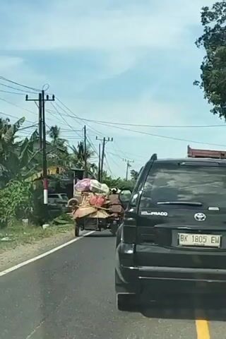 A black Toyota Prado SUV is driving on a paved road. A motorcycle with a sidecar is in front of the SUV, carrying two people and a large amount of cargo. The motorcycle is being driven by a person wearing a brown shirt and a white helmet. The passenger is wearing a patterned shirt and a white helmet. The cargo appears to be household items, including bags, containers, and other miscellaneous objects. The scene is taking place in a rural or suburban area with houses and trees visible on the side of the road. The sky is clear and blue. The license plate on the SUV reads "BK 1805 EW." The motorcycle's license plate is partially visible.