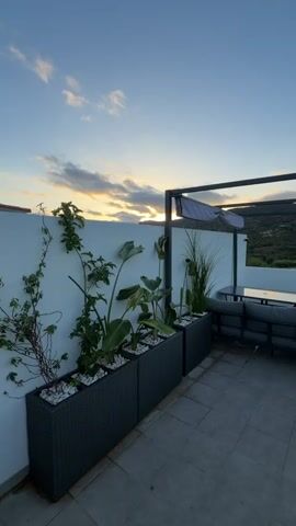 The video pans across a patio area at sunset. In the foreground, there are several long, dark planters filled with white stones and various plants, including some with large, broad leaves and tall grasses. To the right, a white wall forms the backdrop. Further back, a modern dark metal pergola shades an outdoor seating area with a sectional sofa and a table. The background reveals a landscape of rolling hills with green vegetation under a pale blue sky with streaks of orange and yellow clouds. The patio floor is tiled in a light gray square pattern. A glimpse of a dark grill is visible in the lower right corner of the frame.