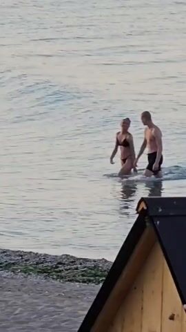 A man and a woman are standing in shallow water near a beach. The woman is wearing a black bikini, and the man is wearing black swim trunks. The man has his arm around the woman, and they are facing each other. The water is calm and clear, and the sky is overcast. There is a small wooden structure in the foreground.