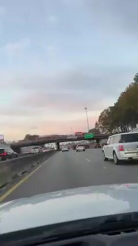 The video shows a car driving on a highway with moderate traffic. An overpass crosses the highway in the distance. A group of people, presumably firefighters, are standing on the overpass. They are wearing white shirts and dark trousers. Two firetrucks are parked on the overpass behind them. The sky above is a mix of pink and gray clouds, suggesting it may be near sunrise or sunset. The camera is positioned from within a vehicle looking at the highway. A sign on the overpass indicates a height restriction of 14 feet 8 inches. The setting appears to be in a suburban or urban environment with trees visible in the background.