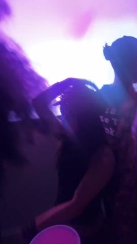 The video shows a group of people at what appears to be a party. The lighting is mostly purple and white, creating a nightclub-like atmosphere. People are dancing and holding red cups. The camera moves around, and the focus changes from one person to another. One individual is wearing a white top.