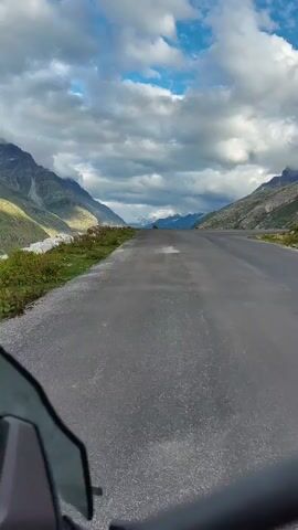 A first-person perspective video captures a motorcycle ride on a paved road through a mountainous valley. The road curves to the right, flanked by green slopes and rocky terrain. Majestic mountains rise on either side, their peaks partially obscured by dramatic, cloudy skies. The sun breaks through the clouds, casting sunlight onto the mountains and illuminating the road ahead. A small portion of the motorcycle's windshield is visible in the lower left corner of the frame. The overall scene conveys a sense of journey and the grandeur of nature.