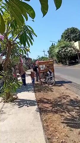 A child is playing on a sidewalk next to a road. A woman is standing nearby, watching the child. A food cart is parked on the side of the road, with a motorcycle next to it. There are trees and houses in the background. The sky is clear and blue.