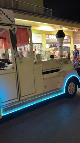 The video shows a tourist train at night, lit with blue and orange lights. The train appears to be white and has a vintage design, resembling an old-fashioned locomotive. The front car has a driver and some passengers. A second car is attached to the back, also carrying passengers. The train is moving along a street lined with shops and restaurants, some of which are also illuminated. Several people are visible on the sidewalk, seemingly watching the train or browsing in the shops. The overall atmosphere is festive and leisurely, suggesting a vacation or tourist destination.