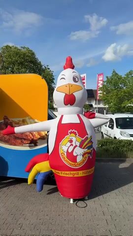 The video shows a large inflatable chicken wearing a red apron with a logo of another chicken on it. The apron has the words "Gaumenschmaus Das Frischgrill-Hähnchen" printed on it. The inflatable chicken has one eye winking and its arms outstretched. It stands on a paved surface next to a yellow food truck with a picture of fried chicken on its side. In the background, there are buildings, trees, flags, and a white car. The sky is blue with some clouds.