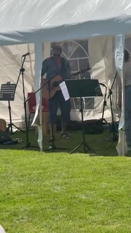 A man plays an acoustic guitar under a white tent. He is wearing sunglasses and a plaid shirt with the sleeves rolled up, and blue shorts. He stands in front of a music stand with sheet music. To his left, another music stand is visible, as are two microphones on stands. To his right, a woman stands, and behind her, a drum set and speakers are partially visible. The ground is covered in green grass, and the tent is set up outdoors. Sunlight illuminates the scene, casting shadows.