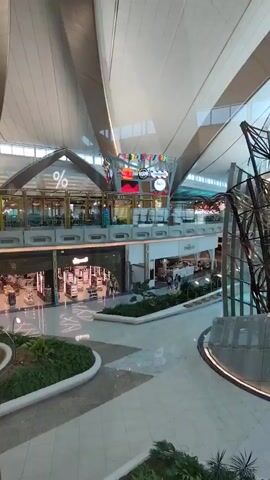The video pans across a large, modern indoor space that appears to be a shopping mall or airport terminal. The architecture is characterized by high ceilings with a complex, multi-layered design, featuring curved white panels and prominent structural beams. Natural light streams in from a large windowed wall on the right side of the frame. In the foreground and midground, there are sections of polished, light-colored flooring with subtle geometric patterns and curves. Landscaping elements, including green bushes and plants, are strategically placed along walkways and in planters.

Several shops are visible on an upper level. One shop sign displays a large percentage symbol (%). Other shops have displays and entrances, with some appearing to be high-end retail establishments. In the center of the midground, there is a large, striking metallic sculpture with a spiraling, cage-like structure, illuminated from below. Digital screens displaying colorful graphics and text are mounted on the upper level, suggesting commercial advertising or information displays.

The space appears clean and well-maintained. There are no people clearly visible in the immediate foreground or midground, but some figures can be seen in the distance within the shop entrances and on the upper walkways, indicating the presence of shoppers or staff. The overall impression is one of a spacious, contemporary, and possibly luxurious environment.