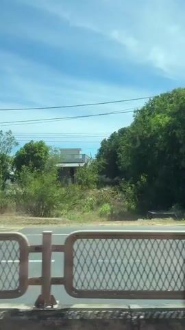 This video captures a scenic view from a moving vehicle, likely a bus or train, traveling through Phong Phú, Vietnam. The camera pans across a landscape bathed in bright sunlight under a clear blue sky with scattered clouds, suggesting a warm, fair-weather day, likely late morning or early afternoon.

The primary subject is the lush, green vegetation lining the roadside, with a prominent focus on large, mature trees. In the foreground, a patterned metal barrier with a diamond-shaped mesh and a few vertical posts suggests the passenger's viewpoint is through a window or opening. Beyond the barrier, a road with white lane markings is visible, along with a sidewalk or paved area.

Interspersed with the natural scenery are glimpses of human infrastructure. A tall, grey utility pole stands to the left, with power lines stretching across the frame. Further along, a small, open-fronted building with a green roof and a sign with Vietnamese text can be seen. Later in the video, a two-story house with a light green facade and a red-tiled roof appears, followed by another building with a red roof. A traffic sign, partially obscured, is also visible.

The overall impression is one of a peaceful, rural or semi-rural journey through a vibrant green landscape under a pleasant sky. There are no people clearly visible, and the focus is on the environment and the passing scenery. The visible Vietnamese text on the building's sign provides a specific location cue, grounding the otherwise generic scenic view in Phong Phú, Vietnam.