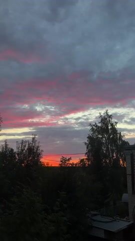 This media file captures a serene sunset scene in Leninsk-Kuznetskiy, Russia. The sky is a dramatic canvas of gray clouds streaked with vibrant pink and orange hues, indicating the final moments of daylight. The setting appears to be a rural or suburban area, with a glimpse of a brick house on the right side of the frame, featuring white-framed windows and a gray, corrugated roof with two chimneys. In the foreground and mid-ground, dense foliage and trees create a silhouette against the colorful sky. A hint of a greenhouse or some agricultural structure is visible in the distance, set against a green field. The overall mood is peaceful and contemplative, evoking the beauty of nature at the end of the day. There is no visible text in the media.