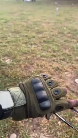 A gloved hand, wearing a tactical style glove that is mostly green with black padded knuckle protectors, holds onto the handlebar of what appears to be a bicycle. The glove also has small black vents on the fingers. A watch-like device is secured to the wrist. The background shows a grassy field or lawn that is slightly uneven with some debris scattered around. In the distance, there is a blurry image of what might be a vehicle and some objects.