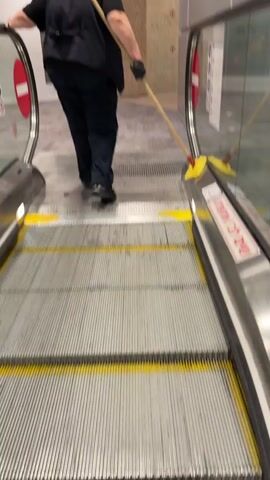 A person wearing a black vest, black pants, and black gloves is mopping the area at the bottom of an escalator. The escalator has yellow safety lines and red and white stop signs on its sides. The floor is made of grey tiles. In the background, there is a fire extinguisher cabinet and a wall made of stone.