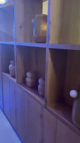 The video shows a long, wooden bookshelf unit with multiple shelves, some of which contain decorative vases and objects. The shelves are illuminated by overhead lights. To the left of the bookshelf, there is a large screen displaying a vibrant underwater scene with fish. The floor is made of polished marble, reflecting the blue ambient lighting and the lights from the bookshelf. In the foreground, part of a couch and two green poufs are visible. The overall environment appears to be a living room or lounge area with a modern and decorative aesthetic.