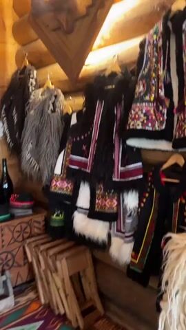 The video shows an interior shot of a room with wooden walls and ceiling. In the foreground, several traditional garments, including vests and ponchos, are displayed on hangers. These garments feature intricate embroidery and fur accents. To the left, a collection of hats and a bottle are visible on a carved wooden chest. Further back, a large tiled stove is prominent, decorated with colorful folk art motifs. Various ceramic bowls and cups are placed on shelves above the stove. A large animal hide hangs on the wall to the right of the stove. The overall impression is of a display of traditional crafts and household items.