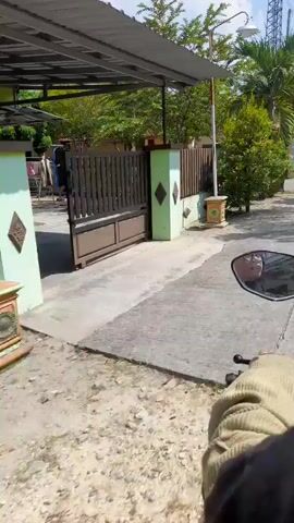A first-person perspective video shows a person riding a motorcycle down a paved street in a village. The rider's gloved hand is visible on the handlebars, and the side mirror reflects the rider's face. The street is lined with houses, fences, and lush green vegetation, including palm trees. The sky is bright blue with scattered clouds. Another motorcyclist is visible in the distance, traveling in the same direction. The video appears to be filmed during the day.