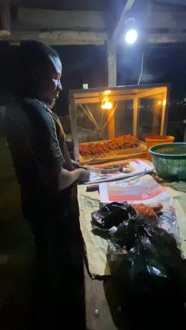 A man is seen standing at a food stall, illuminated by a bright overhead light and the warm glow of a display case filled with skewers of cooked meat. The man, dressed in a dark shirt, appears to be preparing food, handling what looks like paper or leaflets. The stall is set up with a variety of items, including a green basket, an orange container, and black plastic bags filled with something. The surface is covered with newspaper and various items, suggesting a typical food vending setup. Another person is vaguely visible behind the man, partially obscured in the dim lighting. The environment suggests an outdoor or semi-outdoor setting, possibly a street-side stall operating at night.