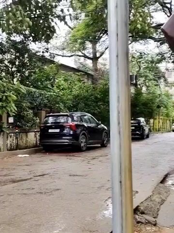 The video shows a street scene. There are parked cars and a motorcycle with a rider. The road appears wet, suggesting it may have rained. Trees and buildings line the street. A metal pole is in the foreground. The motorcycle rider is seen moving away from the camera.