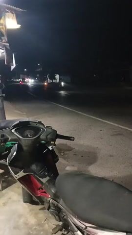 A dark, nighttime scene shows a street with a motorcycle in the foreground. The motorcycle is dark-colored with red accents and is parked on a paved surface. The street is wet, indicating recent rain or damp conditions. Headlights from oncoming vehicles illuminate the wet asphalt and create reflections. In the background, a few figures can be seen, possibly pedestrians or other vehicles, moving along the street. Streetlights and building lights are visible, casting a dim glow on the environment. The overall atmosphere is quiet and still, with the sounds of passing vehicles being the most prominent audible elements.