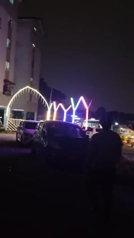 The video shows a nighttime scene with artificial lights illuminating an outdoor area. In the foreground, the image is dark and blurry, with hints of greenery and possibly a car. Further back, a row of brightly lit structures is visible, possibly stalls or small buildings. These structures are decorated with colorful lights, including a prominent triangular design with purple and green lights. People are seated at tables in front of these structures, suggesting an eating or gathering area. In the background, tall buildings with many illuminated windows are visible, indicating an urban or suburban setting. The overall atmosphere is lively due to the lights and activity.