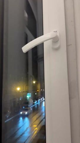 A hand opens a white window with a handle labeled “SOLO” to reveal a nighttime street scene. The street is wet, reflecting the lights of passing cars and streetlights. Buildings line the street, and two people can be seen walking on the sidewalk. The sky is dark.