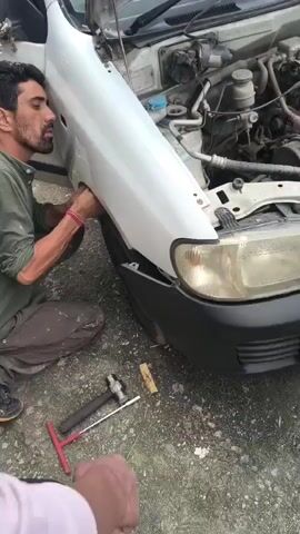 A man is kneeling on the ground next to a white car, working on the fender near the headlight. He is wearing a green shirt and dark pants. Tools, including a hammer and a T-handle screwdriver, are on the ground near him. Another person is visible in the foreground, wearing white slides. The scene appears to be outdoors, with a rough, grey surface.