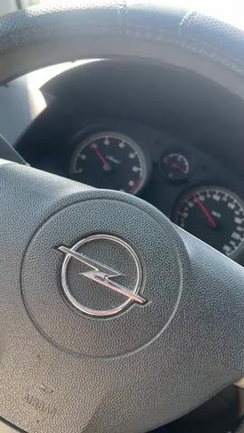 The video shows the interior of a car, focusing on the steering wheel and dashboard. The car is in motion, indicated by the moving speedometer and the view of the road ahead through the windshield. The steering wheel has the Opel logo in the center. The dashboard displays several gauges, including a speedometer with the needle indicating a speed of approximately 100 km/h, and a tachometer. The external environment is a clear, sunny day with scattered clouds, and there are trees, buildings, and other vehicles visible on the side of the road. A blue bus is seen ahead of the car, traveling in the same direction. The overall impression is of a car driving on a highway or a similar road under good weather conditions.