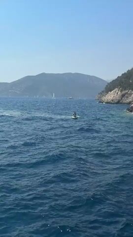 A jet ski and a sailboat are visible on the bright blue sea. The camera pans right to show a rocky coastline with trees and caves. A person's hands are visible holding a smartphone, presumably taking a picture of the caves. The water is clear and turquoise near the shore.