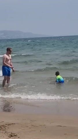 A man in blue swim trunks stands in shallow ocean water, facing away from the camera, towards the right. A child in a green and blue swimsuit is in the water to the right of the man, also facing away from the camera. The child is in a crouched position, appearing to be playing in the waves. The man raises his right hand to his face, possibly adjusting sunglasses or shielding his eyes. The ocean has small waves breaking on a sandy beach. In the background, a hazy coastline with hills is visible under a bright sky.