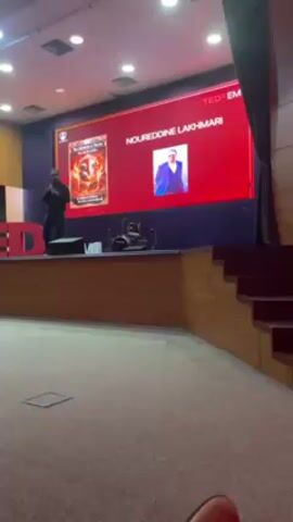A man in a black shirt is standing on a stage giving a TEDx talk. A large screen behind him displays his name, Noureddine Lakhmari, along with a headshot and an image of a book cover. He gestures with his hands while speaking. The stage is made of wood. The seating area in front of the stage is carpeted. There are stairs to the right of the stage. The walls of the auditorium are painted blue.