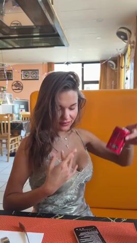 A young woman with long, brown hair is sitting at a table in a restaurant. She is wearing a sparkly, low-cut dress with lace trim. She is holding a red soda can in her right hand and looking at it. Her left hand is raised, and she is touching her face. The restaurant has orange booths and tables with orange placemats. There is a phone on the table.
