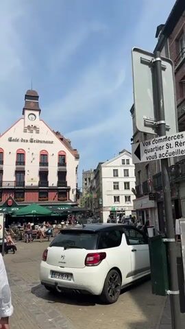 The video shows a street scene with pedestrians and vehicles on a sunny day. In the foreground, a white car is parked. Across the street, a cafe called "Cafe des Tribunaux" is visible, with outdoor seating occupied by patrons. The architecture of the buildings suggests a European setting. People are walking on the sidewalk, some are sitting at the cafe, and others are in cars. A signpost with French text and an arrow indicates directions. The sky is blue with some clouds. The camera pans and moves to capture different aspects of the street.