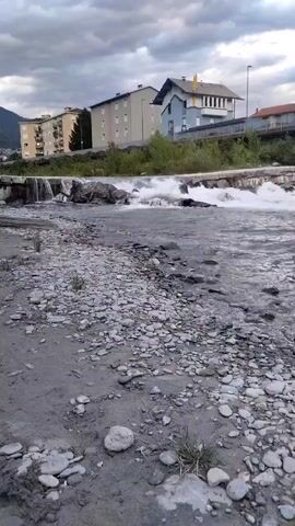 The video shows a wide shot of a river flowing past a rocky bank. Buildings are visible on the opposite bank, with trees and a road in between. The water is turbulent, with small waterfalls visible. The sky is cloudy. The camera is positioned at a low angle, looking across the river.