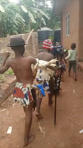 Several children walk along a path outside a building. They are bare-chested and wear patterned fabric tied around their waists. One child has a fedora hat. Another carries a short white fringe that appears to be made of straw or grass. Another child carries a walking stick. A child in a green shirt runs ahead of them. Water tanks are visible in the background. The path is dirt.