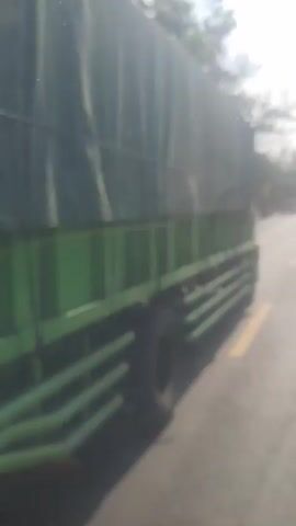 The video is a blurry, first-person perspective of a person traveling on a road. A large green truck is visible on the left side of the screen, taking up a significant portion of the frame. The truck appears to be in motion, indicated by the slight blur. The road itself is paved and has a yellow line marking the lane. In the distance, other vehicles can be seen, also appearing blurry. The environment seems to be rural, with trees and vegetation visible on the sides of the road. The sky is bright and overcast, suggesting daylight. The overall impression is of a moving vehicle traveling along a highway, with a truck as a prominent foreground element.