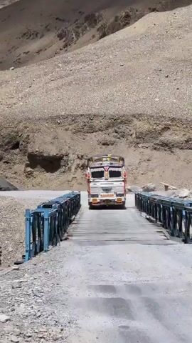 A truck is driving across a narrow, wooden bridge. The bridge has blue railings. The road leading to the bridge is unpaved and rocky. The truck is white with colorful decorations on the front and top. The background is a rocky, barren mountainside. The sky is overcast.
