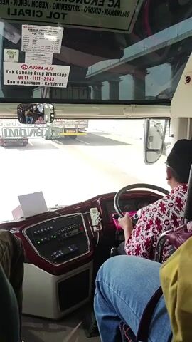 The video shows the interior of a bus, looking forward from a passenger's seat. A driver, wearing a black head covering and a red and white patterned shirt, is seated at the controls, holding the steering wheel. The dashboard features various buttons and a radio. Through the windshield, other vehicles, including trucks and buses, are visible on a road under an overpass. A sign on the windshield reads "PRIMA JASA" and "Yuk Gabung Grup WhatsApp 0811-1111-2343 Route Kuningan - Kalias". The scene appears to be in motion, suggesting the bus is traveling.