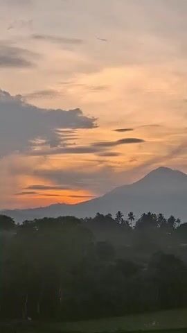 The video shows a sunset over a landscape. The sky is filled with orange and yellow clouds, and the sun is setting behind a mountain. In the foreground, there are trees and rice paddies. The overall scene is peaceful and serene.