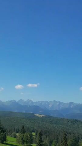 A drone shot pans across a mountain landscape. In the foreground, there is a dense forest. Behind the forest, there are rolling hills and a range of majestic mountains. The sky is a vibrant blue with a few scattered clouds. The drone then moves forward, revealing the roof and side window of a car.