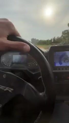 A first-person perspective shot shows a person driving a Polaris RZR on a dirt road. The sun shines brightly in the sky. The driver’s hand is visible on the steering wheel. The dashboard displays a speedometer that registers increasing speed, reaching 82 mph. The terrain appears to be rural, with sparse vegetation and buildings on the side of the road.