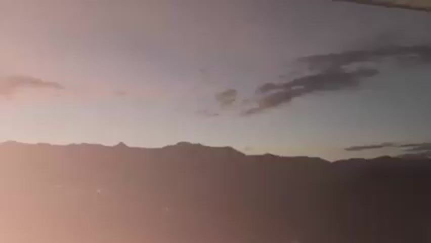 The video shows a wide shot of a mountain range silhouette against a twilight sky with scattered clouds. The sky has gradients of blue, purple, and pink hues. There's a faint glow from a town at the base of the mountains. A light source appears to come from the lower-left corner of the frame, creating a lens flare effect. A white, horizontal bar is visible at the top edge of the frame. Music plays in the background.